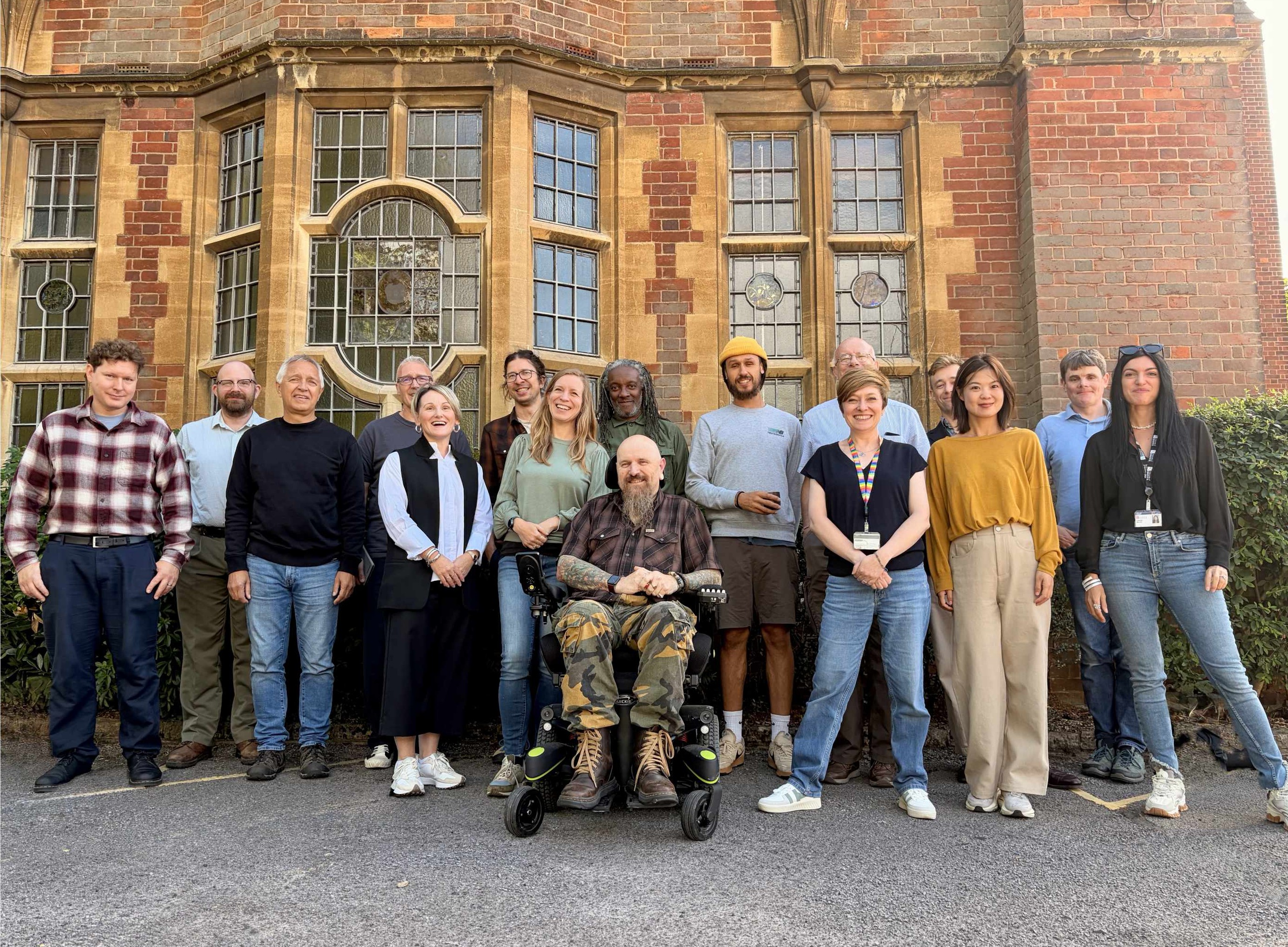 Group portrait of 15 people standing outdoors in front of a large historic red-brick building with tall, decorative leaded windows and stone surrounds. One person is seated in a powered wheelchair at the centre of the front row, with the rest of the group standing closely together on either side.